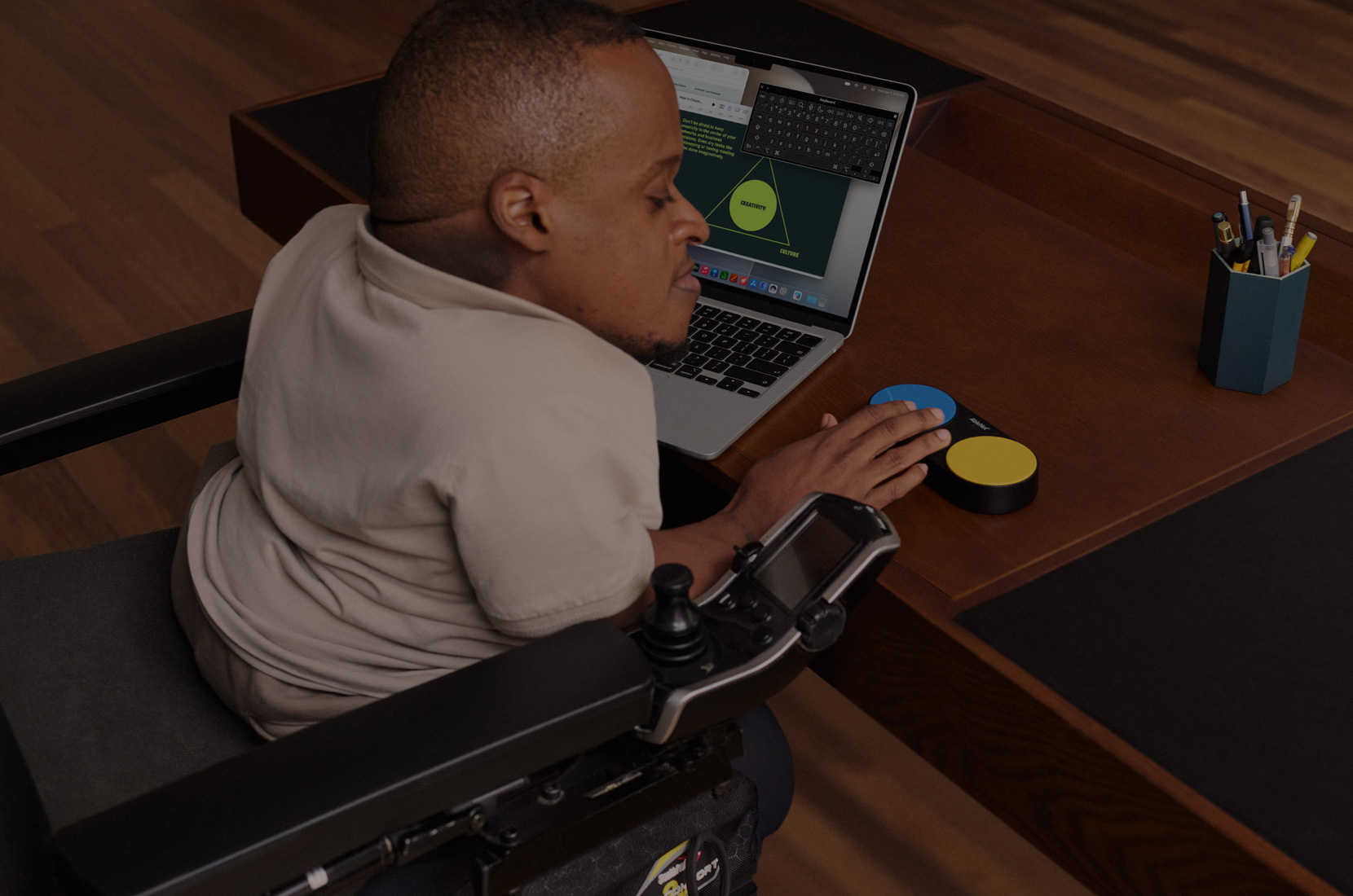 Foto de un hombre en una silla de ruedas eléctrica en un escritorio usando la funcionalidad Teclado de Accesibilidad en una MacBook Air de 13 pulgadas y un dispositivo con Control por Botón