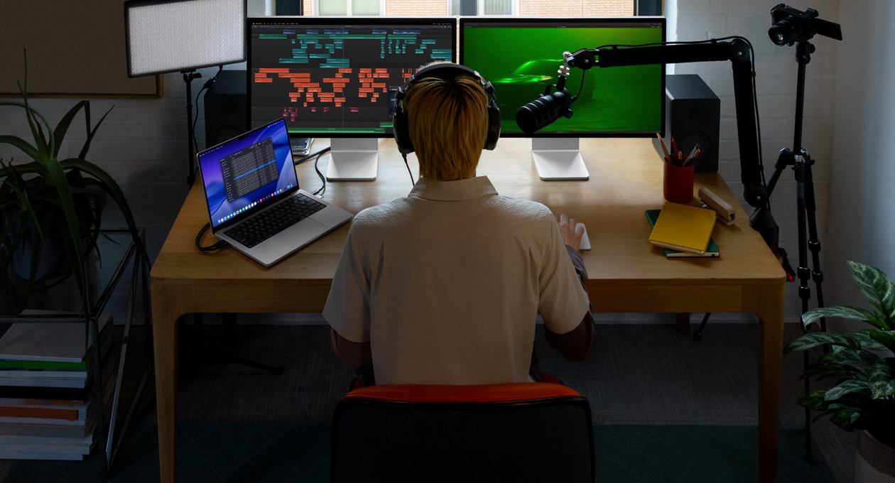 Persona sentada en un escritorio con un micrófono y audífonos frente a tres dispositivos: un monitor Studio Display para la Mac, herramientas de edición de audio, clips de sonido en diferentes bloques de color, un segundo monitor Studio Display para la Mac, foto de un carro en la niebla con iluminación ambiental verde, y una MacBook Pro de 16 pulgadas con la app Finder abierta, Archivos para Final Cut Pro, el Dock y un fondo de escritorio azul estilizado
