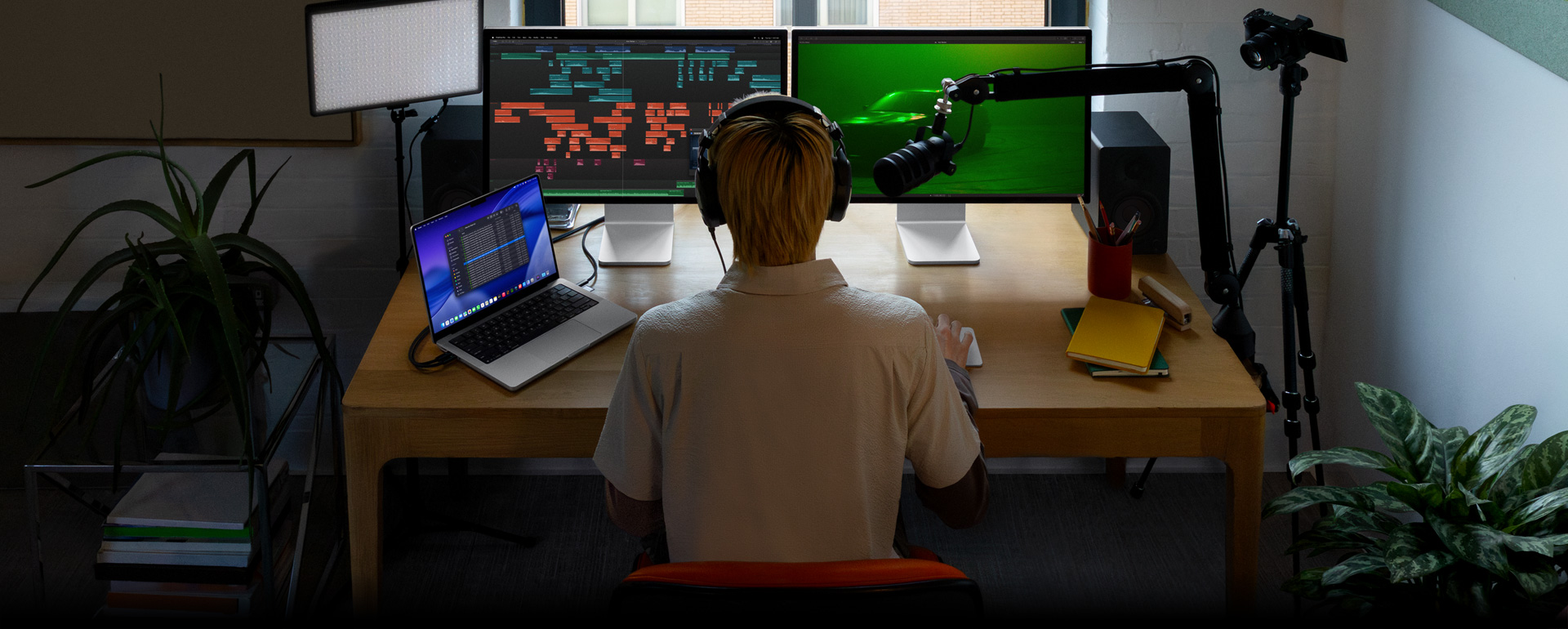 Persona sentada en un escritorio con un micrófono y audífonos frente a tres dispositivos: un monitor Studio Display para la Mac que muestra herramientas de edición de audio y clips de sonido en distintos bloques de color, un segundo monitor Studio Display para la Mac que muestra un carro en una escena con iluminación ambiental verde, y una MacBook Pro de 16 pulgadas con una larga lista de tomas en un proyecto de Final Cut Pro