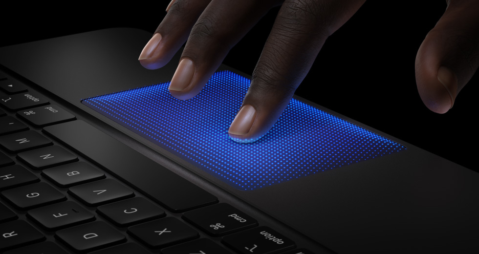 Un Magic Keyboard, parte lateral, color negro, una mano pasa por encima del trackpad, el dedo índice con una sombra azul muestra cómo funciona