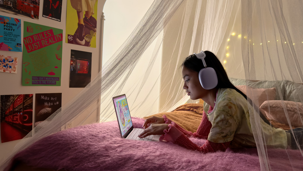 Una joven en su habitación con audífonos puestos usa una MacBook Neo, rosa rubor, para agendar una cita de manicure