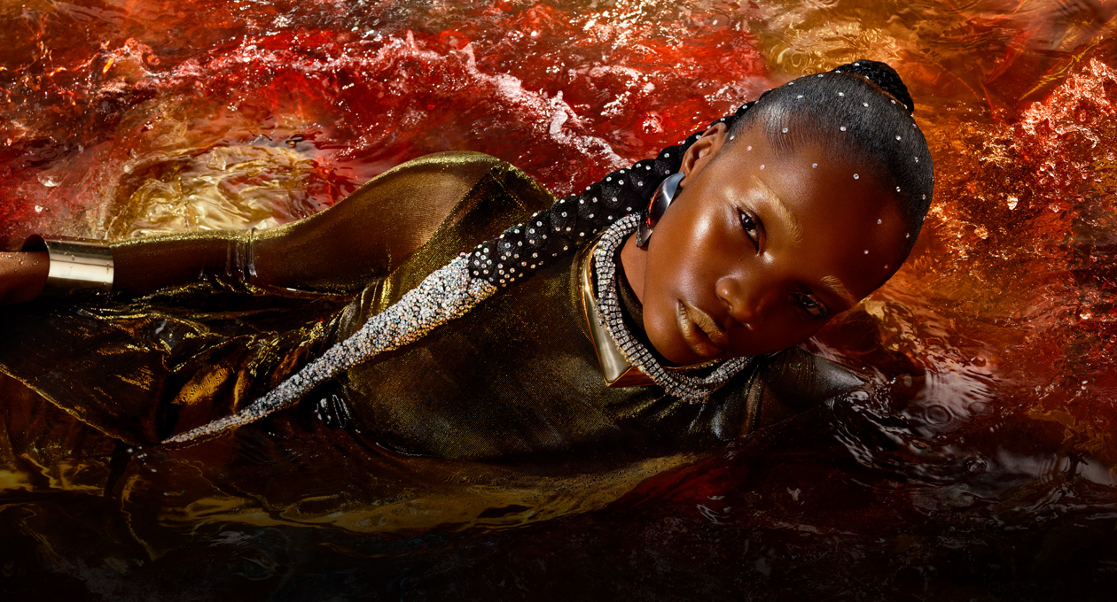 Una modelo con un vestido color oro metálico acomodada de lado en medio de un cuerpo de agua rojo, se muestra el brillo, el contraste y la amplia gama de colores del monitor Studio Display XDR